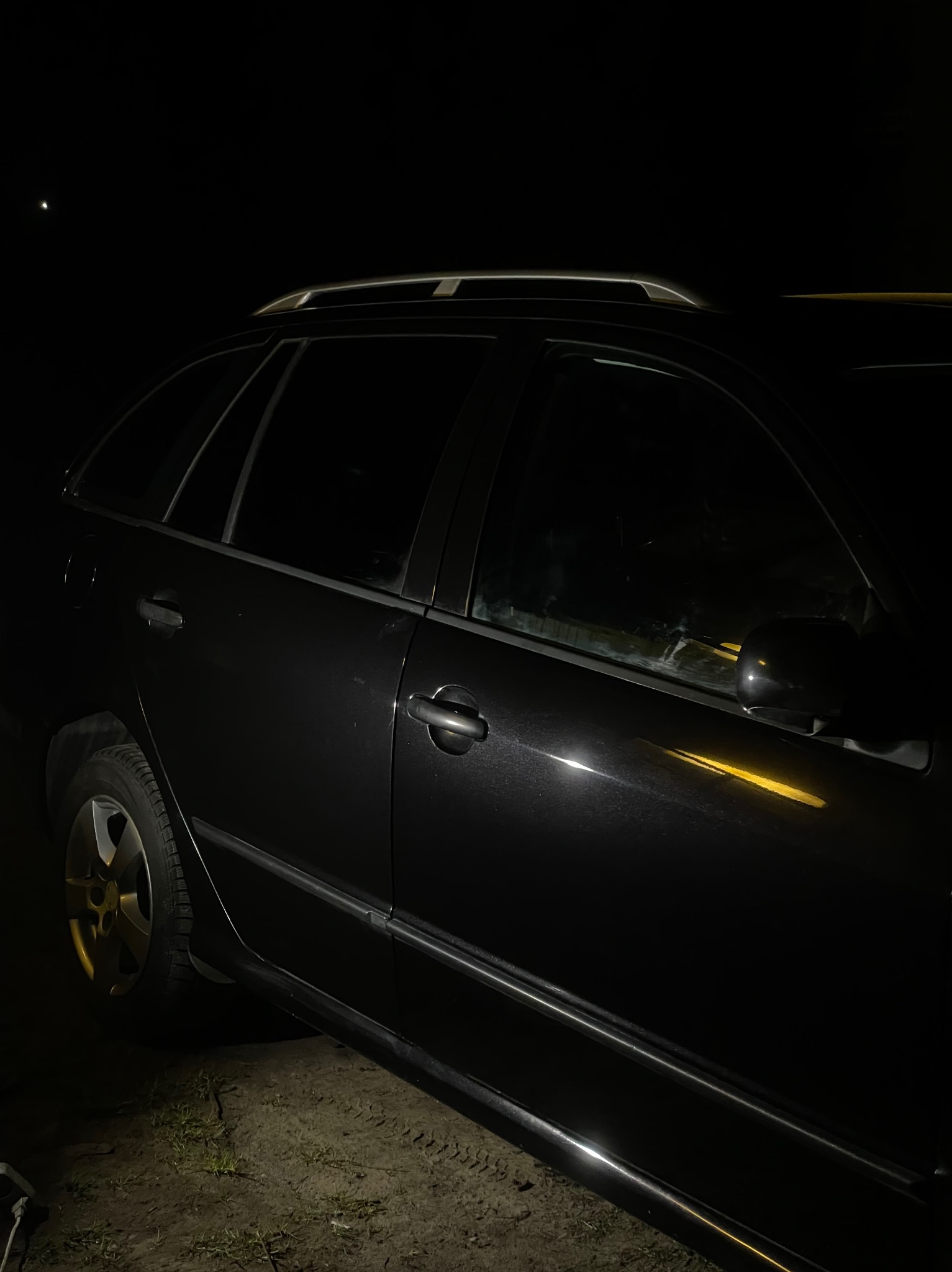 Ciemne zdjęcie boku czarnego kombi zaparkowanego na ziemi, z widocznymi oponami i relingami dachowymi.