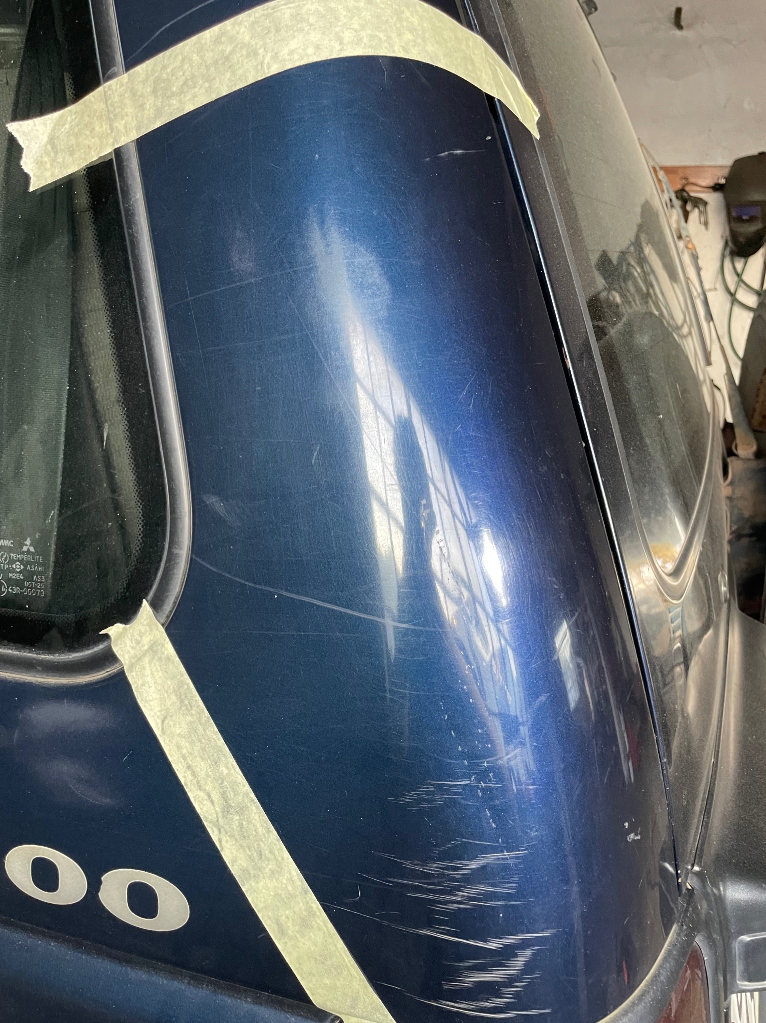 Fragment karoserii granatowego samochodu z widocznymi zarysowaniami, zabezpieczony taśmą malarską przed naprawą, w tle warsztat.