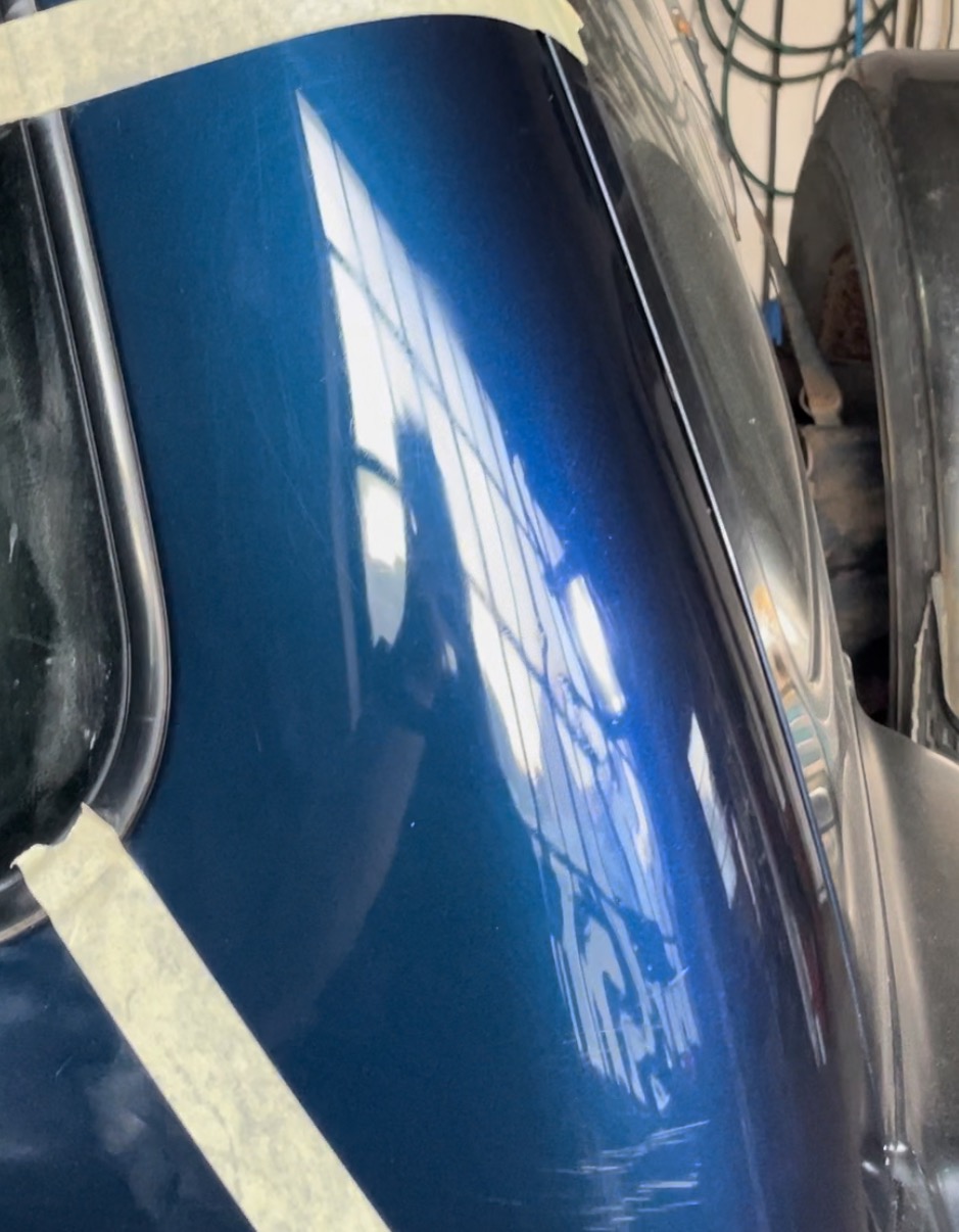 Błyszczący, niebieski fragment karoserii samochodu z widocznymi zarysowaniami i taśmą maskującą, odbijający jasne okno.