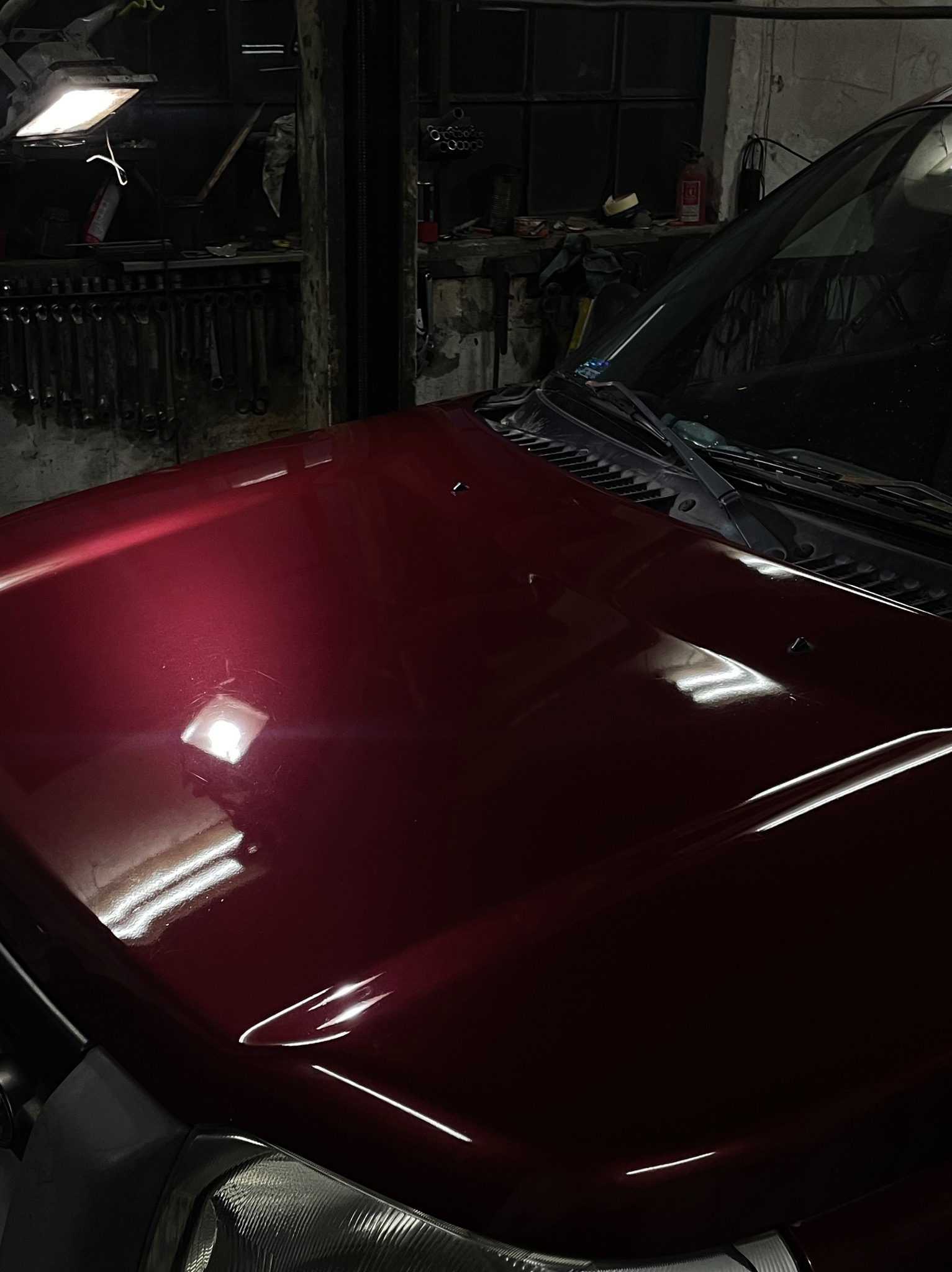 Bordowy lakierowany błotnik samochodu z widoczną lampą i szybą, na tle warsztatu z narzędziami.