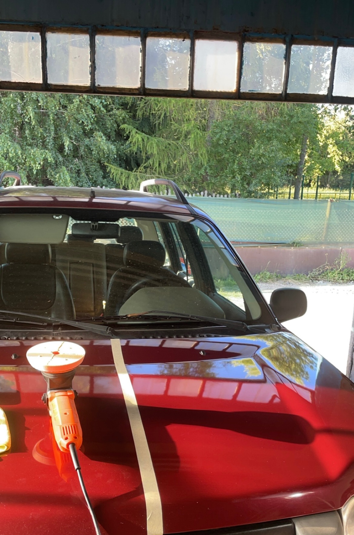 Polerka na masce bordowego samochodu, widoczny efekt przed i po polerowaniu, tło z zielenią przez okno warsztatu.