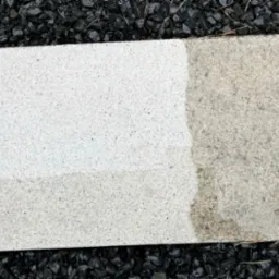 Kostka brukowa częściowo umyta, ukazująca różnicę między czystą a brudną powierzchnią, otoczona czarnym grysem i fragmentem trawy.