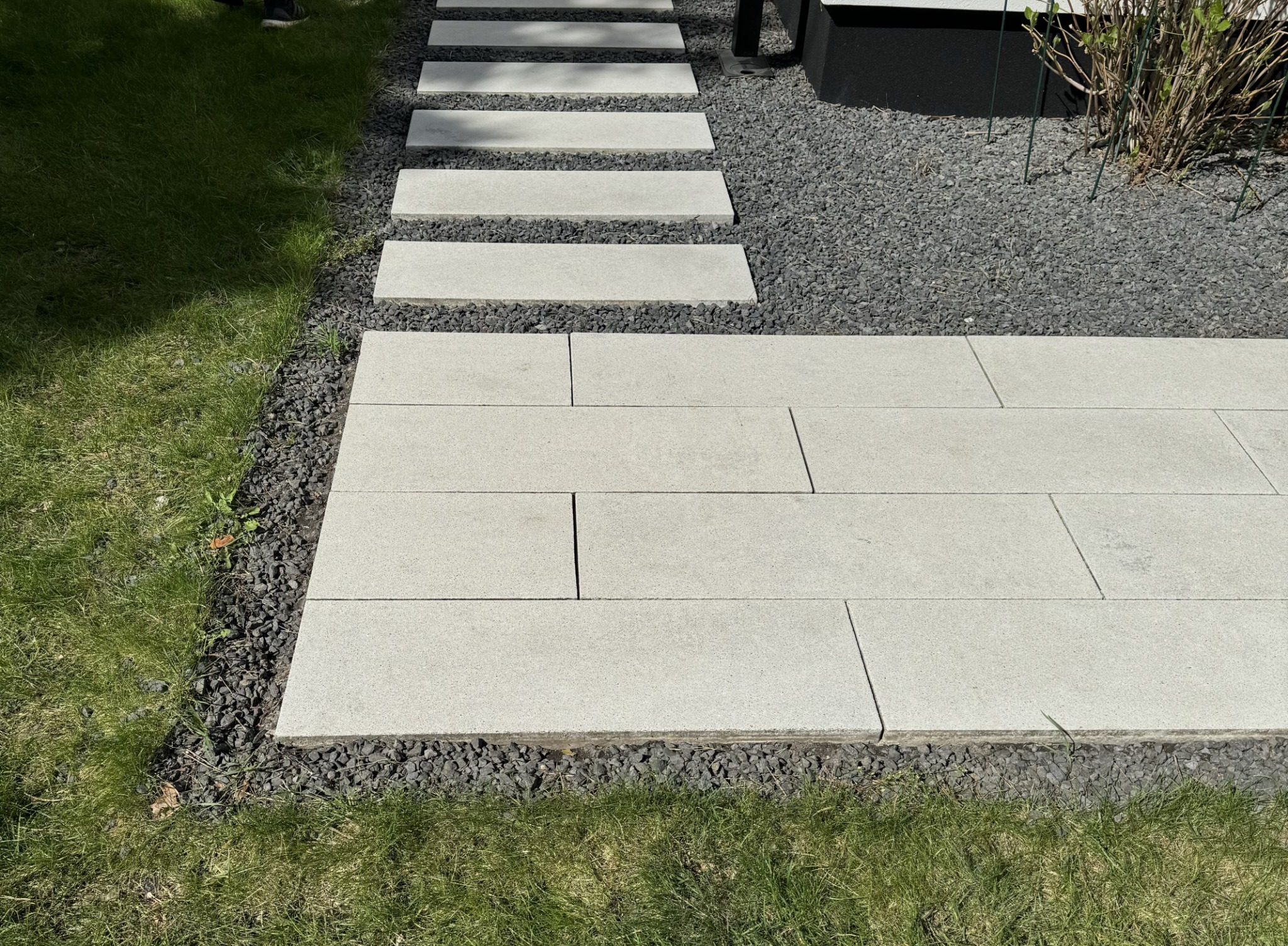 Ułożona ścieżka z jasnych płyt chodnikowych i prostokątna powierzchnia z kostki brukowej otoczona czarnym grysem i trawnikiem.