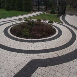 Tomasz Brilowski - Okrągły wzór z jasnoszarej i ciemnoszarej kostki granitowej tworzący ścieżkę i podjazd, z centralnym rabatem roślinnym, zakończony kutą bramą w tle.