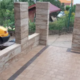 Wykończony taras z kostki brukowej w odcieniach beżu z ciemnobrązową obwódką, otoczony słupkami z tego samego materiału, z widocznym żółtym samochodem w tle.