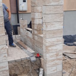 Tomasz Brilowski - Wznoszenie murku z bloczków imitujących trawertyn, widoczne fragmenty chodnika z kostki brukowej, robotnik z łopatą oraz elementy instalacji elektrycznej na podłożu.