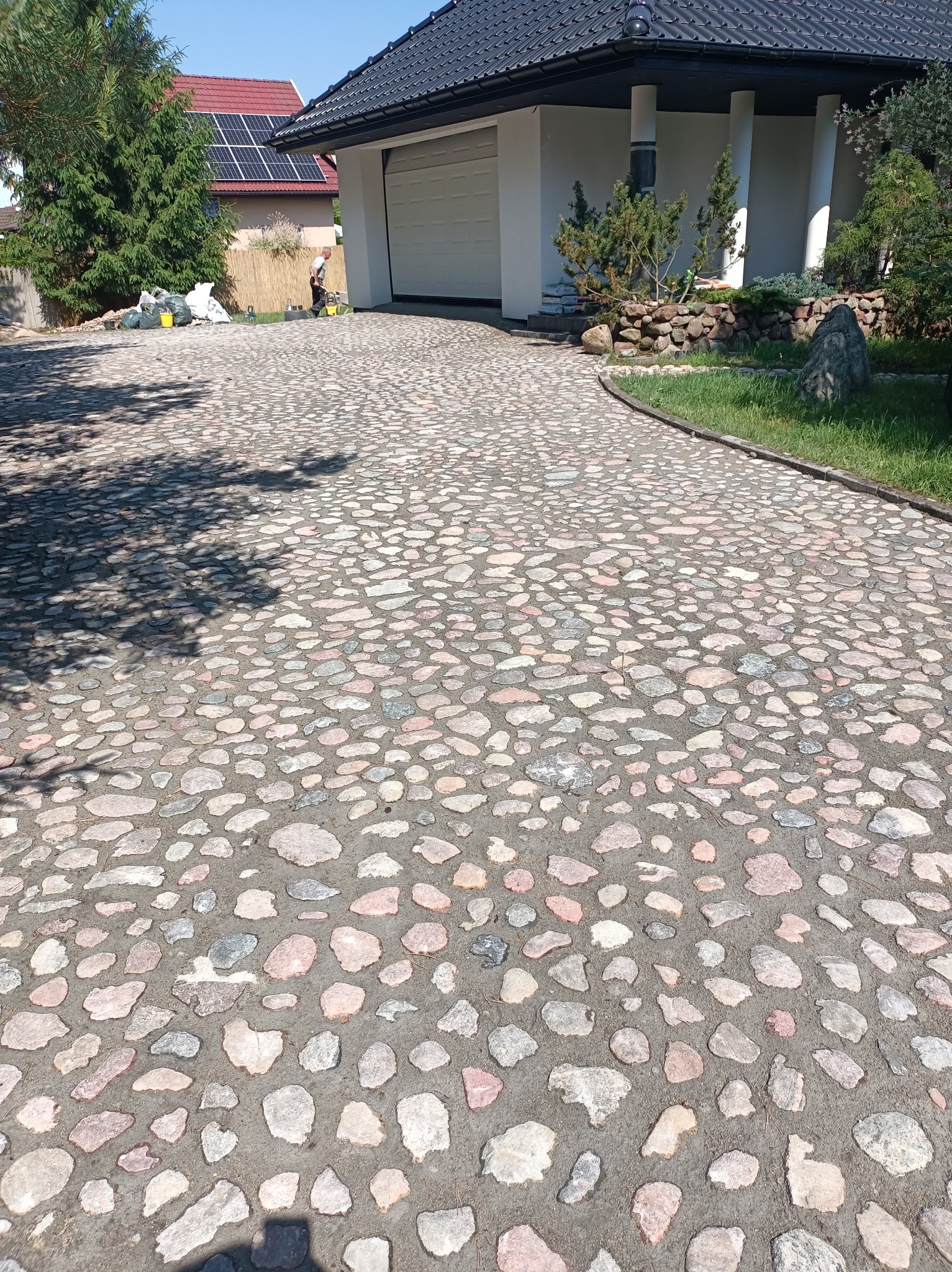 Podjazd wyłożony nieregularną kostką granitową w odcieniach szarości i różu, prowadzący do białego domu z garażem; w tle widoczna osoba pracująca przy ogrodzie.