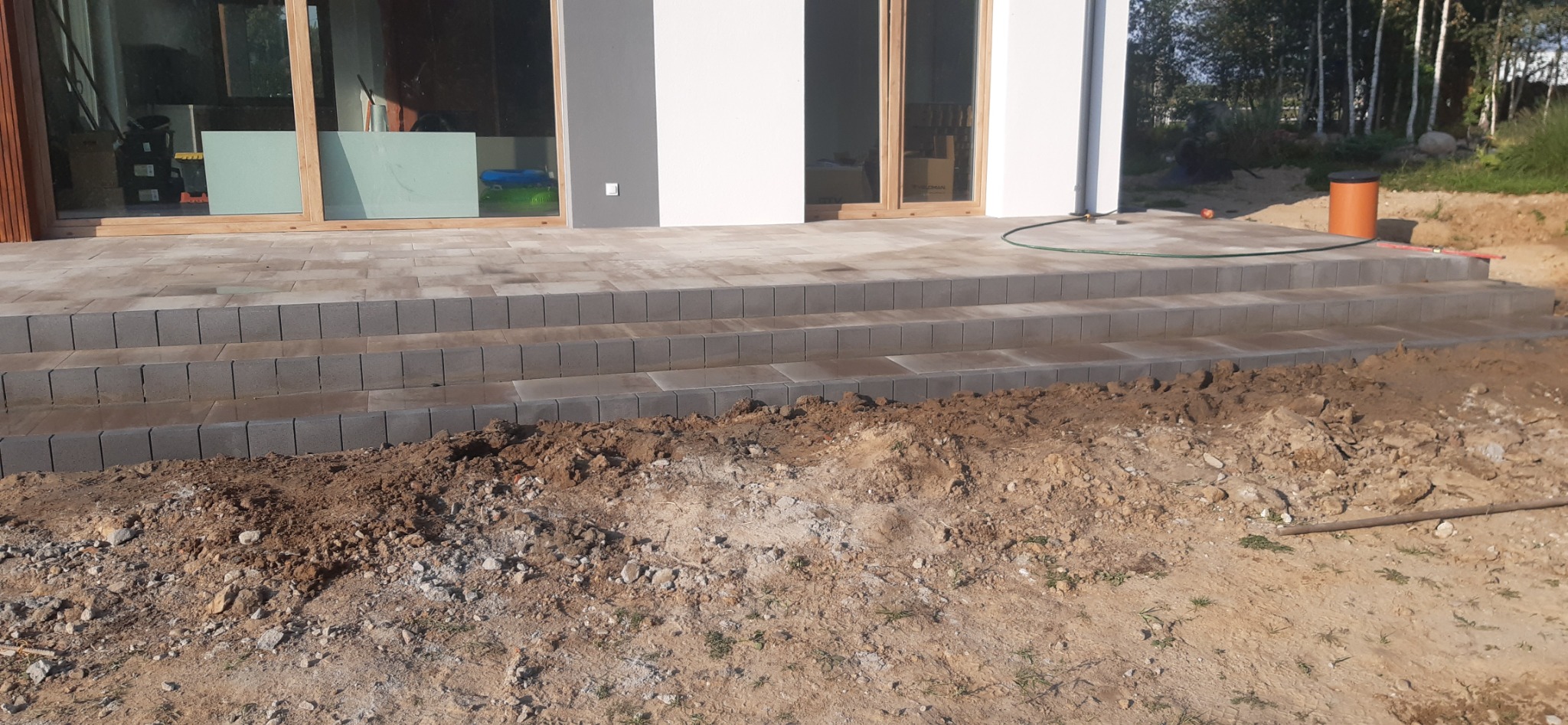 Nowe, szare schody z kostki brukowej prowadzące do nowoczesnego domu z dużymi, przesuwnymi drzwiami tarasowymi; widoczne ślady prac ziemnych wokół schodów.