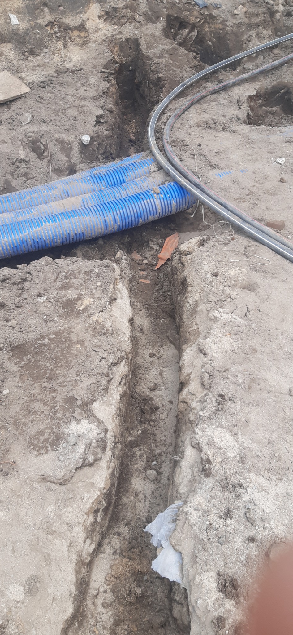 Wykopany wąski rów w ziemi, widoczne niebieskie rury karbowane i czarne kable ułożone obok rowu, fragment cegły i kawałek plastikowej folii w wykopie.