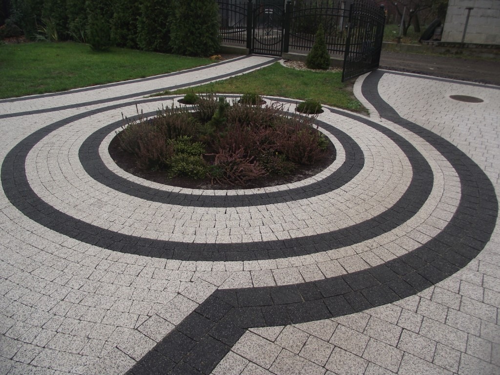 Okrągły wzór z jasnoszarej i ciemnoszarej kostki granitowej tworzący ścieżkę i podjazd, z centralnym rabatem roślinnym, zakończony kutą bramą w tle.