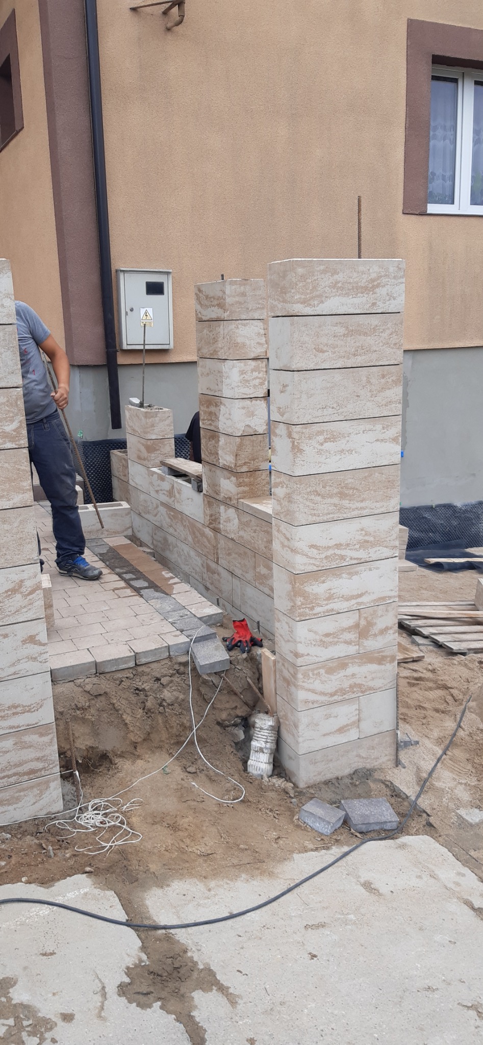 Wznoszenie murku z bloczków imitujących trawertyn, widoczne fragmenty chodnika z kostki brukowej, robotnik z łopatą oraz elementy instalacji elektrycznej na podłożu.