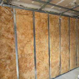 RóżMik - Montaż izolacji z wełny mineralnej w konstrukcji metalowej ściany działowej z widocznymi przewodami elektrycznymi w tle surowy sufit z niedokończonymi pracami gipsowymi.