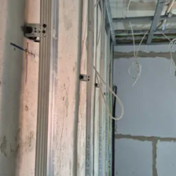 Montaż profili metalowych do ściany z bloczków, widoczne przewody elektryczne, konstrukcja sufitu podwieszanego w tle.