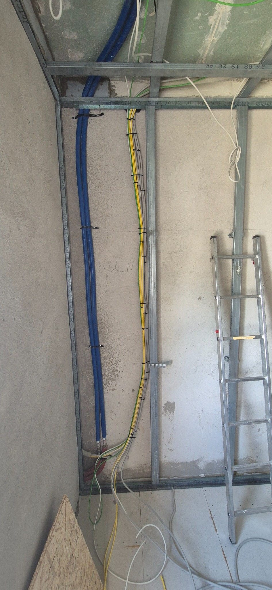 Wnętrze w trakcie remontu: widoczne metalowe profile konstrukcyjne, pionowo poprowadzone niebieskie rury i wiązka kabli elektrycznych w żółto-zielonych osłonach, przymocowane do ściany za pomocą...