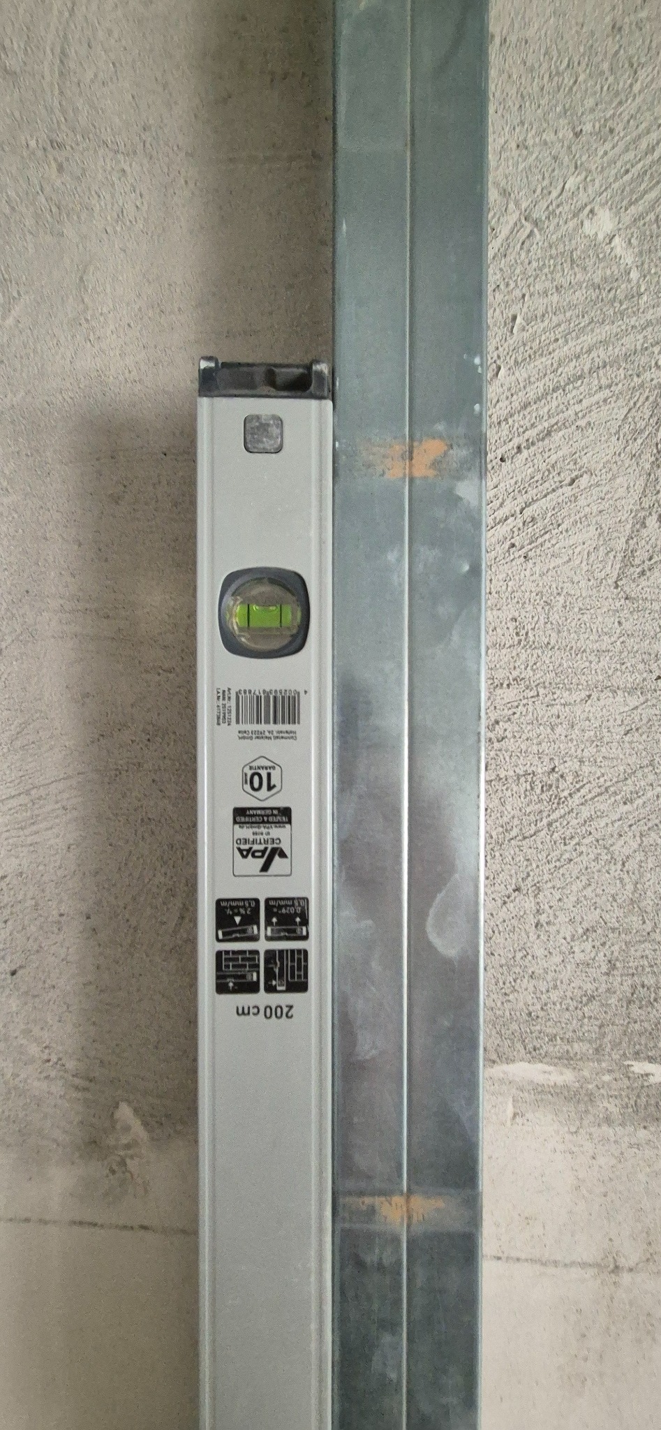 Poziomnica przyłożona do metalowego profilu na tle surowej ściany, prawdopodobnie podczas montażu konstrukcji z płyt kartonowo-gipsowych.