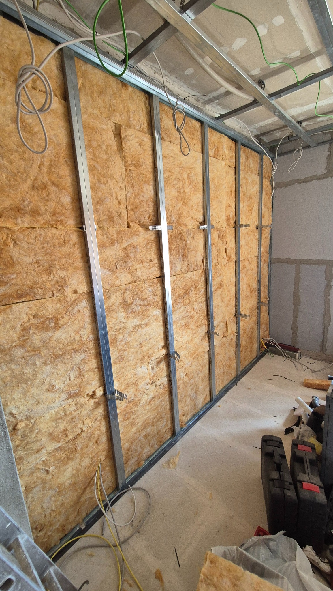 Montaż izolacji z wełny mineralnej w metalowym stelażu na ścianie, widoczne przewody elektryczne i narzędzia na podłodze.