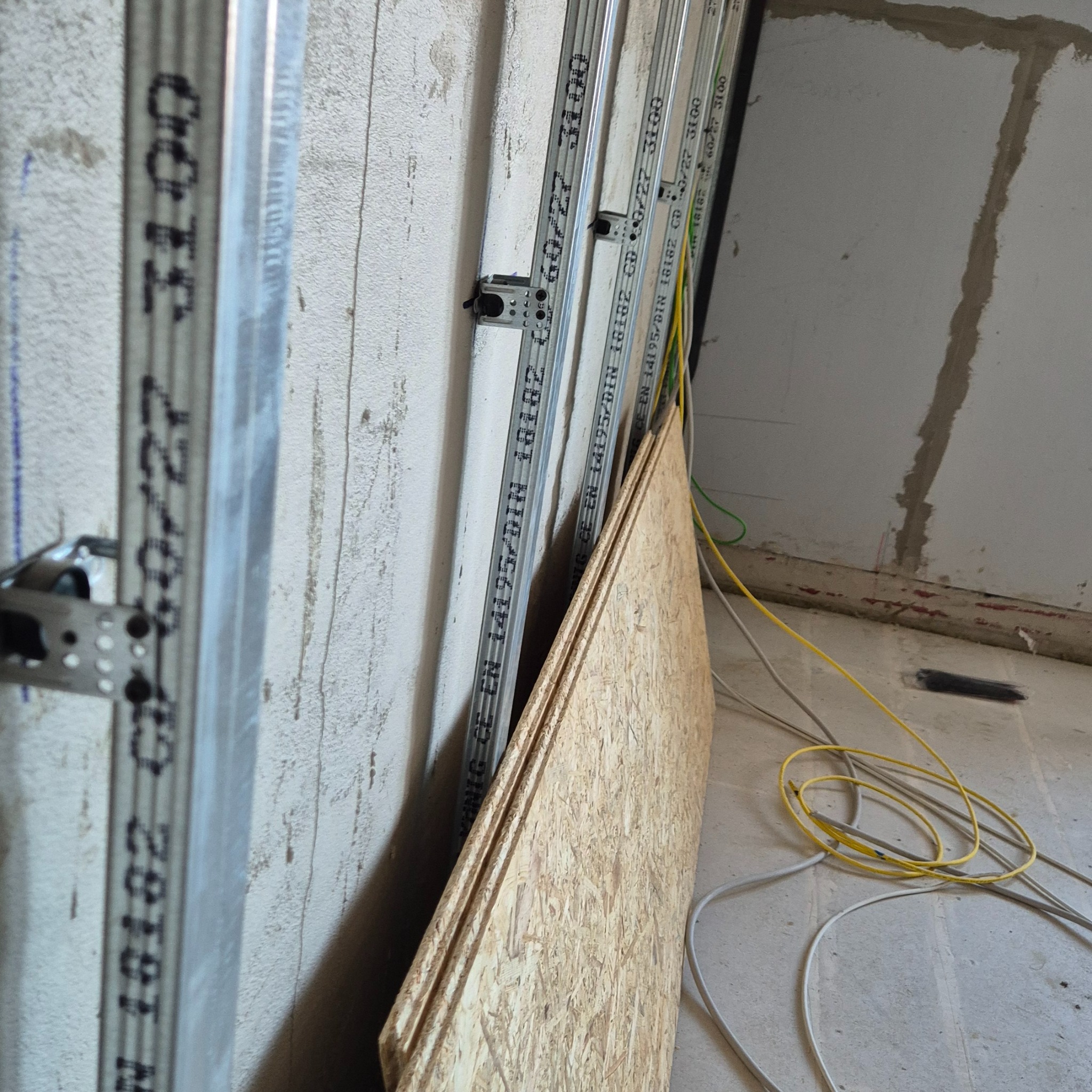 Stojące profile metalowe do płyt kartonowo-gipsowych z oznaczeniami producenta, obok płyty OSB i luźno leżące przewody elektryczne na podłodze w surowym stanie.