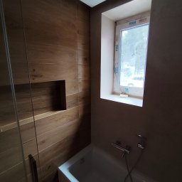 Kamil Kiszka - Wykończona łazienka z wanną, prysznicem za szklaną ścianką i drewnopodobnymi płytkami na ścianach, wnęka dekoracyjna w ścianie, okno z widokiem na zewnątrz.