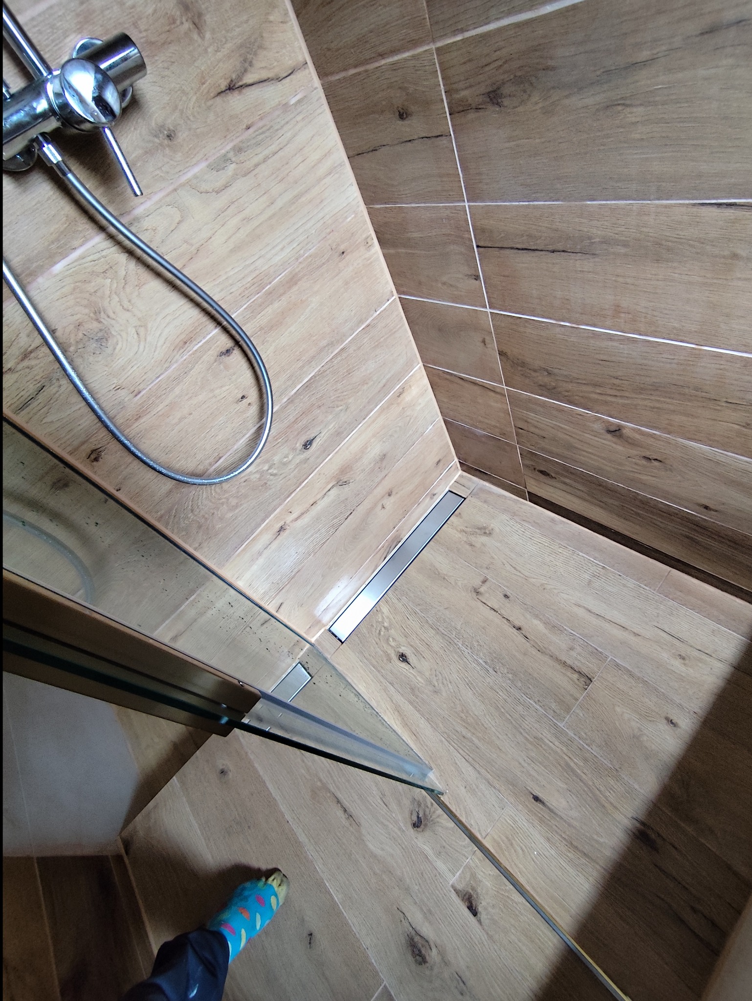 Wykończona kabina prysznicowa walk-in z odpływem liniowym i armaturą w kolorze chromu, płytki imitujące drewno na ścianach i podłodze, realizacja w Gliwicach, widoczna stopa w kolorowej skarpetce.