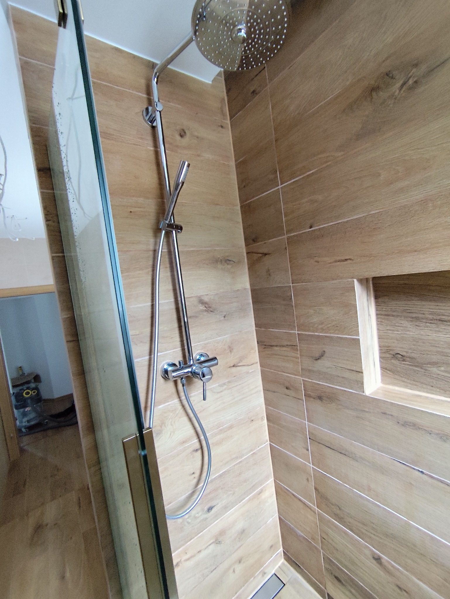 Nowoczesna kabina prysznicowa z deszczownicą i panelami ściennymi imitującymi drewno, widoczny fragment szklanej ścianki i półka w ścianie.