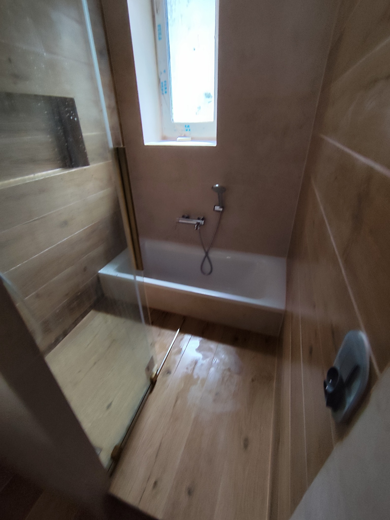 Niewielka łazienka na poddaszu z wanną, prysznicem i drewnopodobnymi płytkami na ścianach i podłodze, widok zza szklanej kabiny.