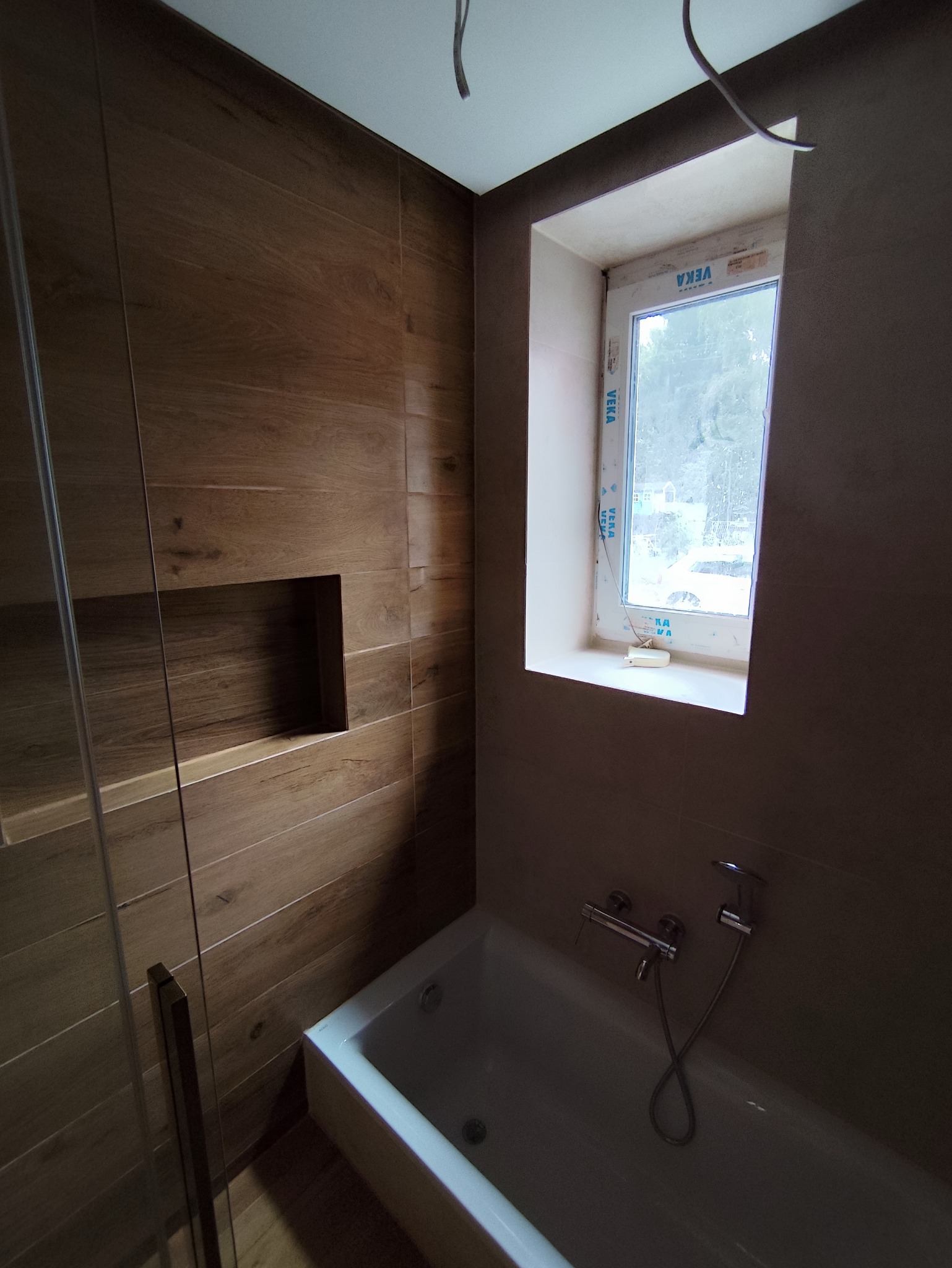 Wykończona łazienka z wanną, prysznicem za szklaną ścianką i drewnopodobnymi płytkami na ścianach, wnęka dekoracyjna w ścianie, okno z widokiem na zewnątrz.