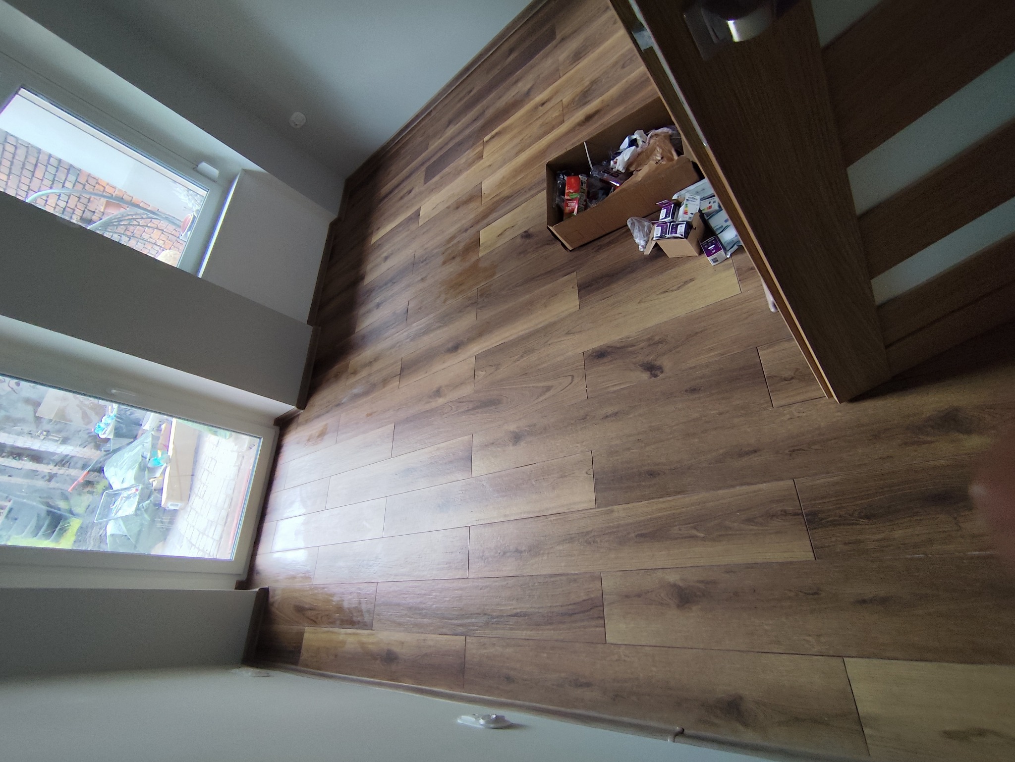 Ułożone panele podłogowe w odcieniach brązu, widoczne niedokładności montażu przy ościeżnicy drzwi oraz karton z pozostałościami po materiałach budowlanych.