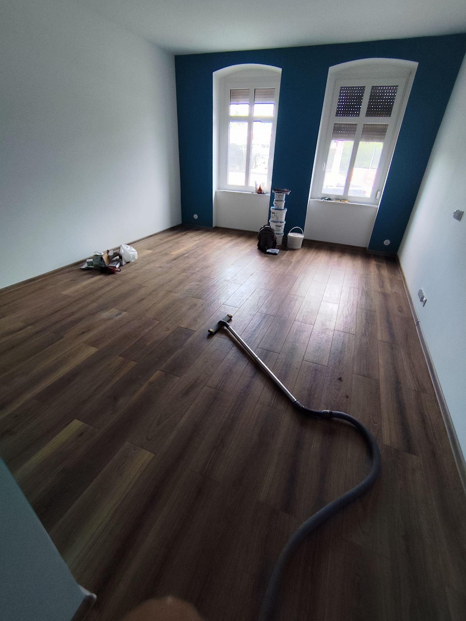 Pokój po odświeżeniu z nową, ciemną podłogą panelową, świeżo pomalowaną niebieską ścianą z dwoma oknami i białą ścianą boczną, widoczne puszki po farbie oraz odkurzacz do podłóg.