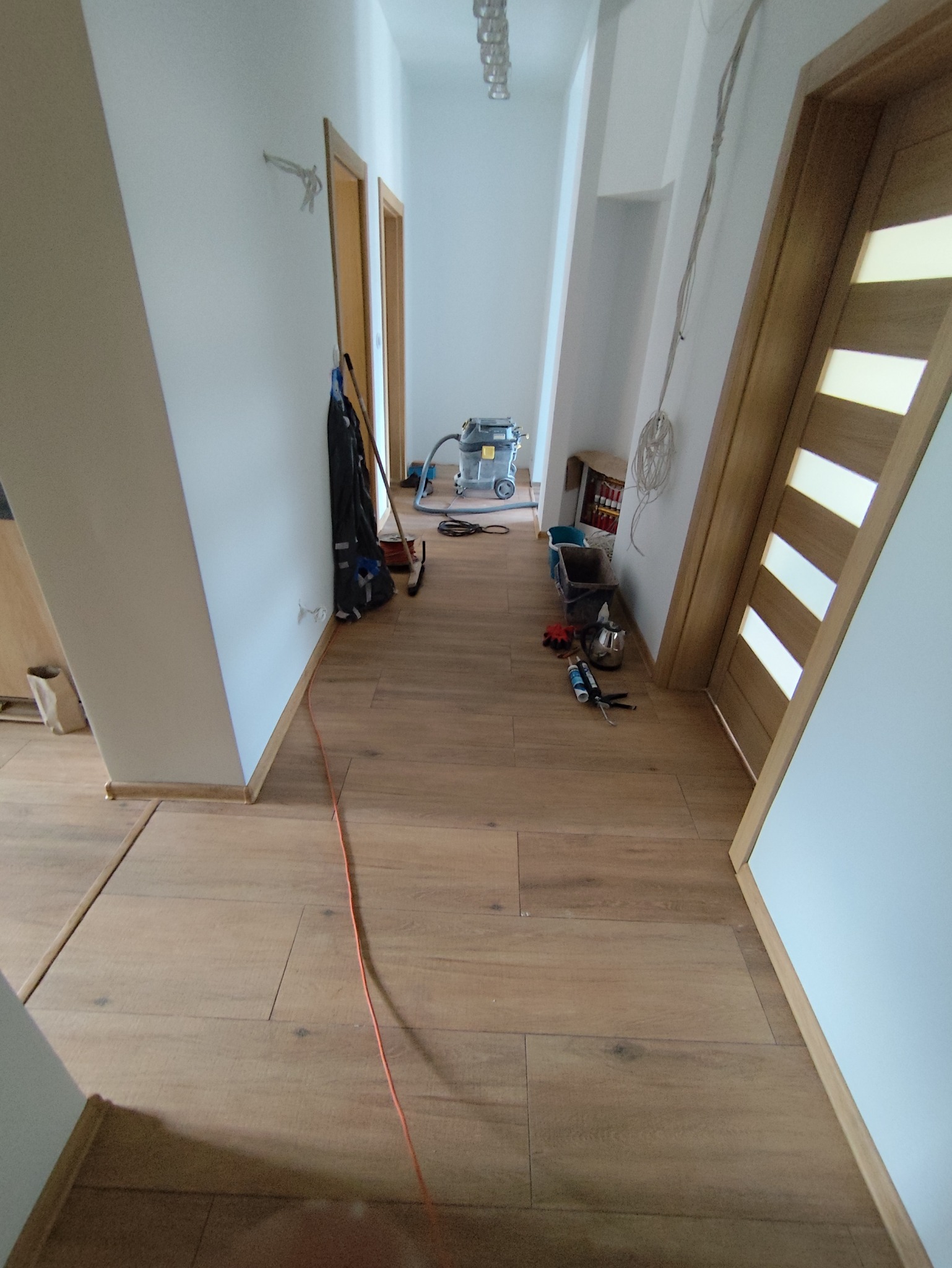 Korytarz w trakcie prac wykończeniowych z widocznymi narzędziami, odkurzaczem przemysłowym i częściowo położoną podłogą z paneli imitujących drewno.