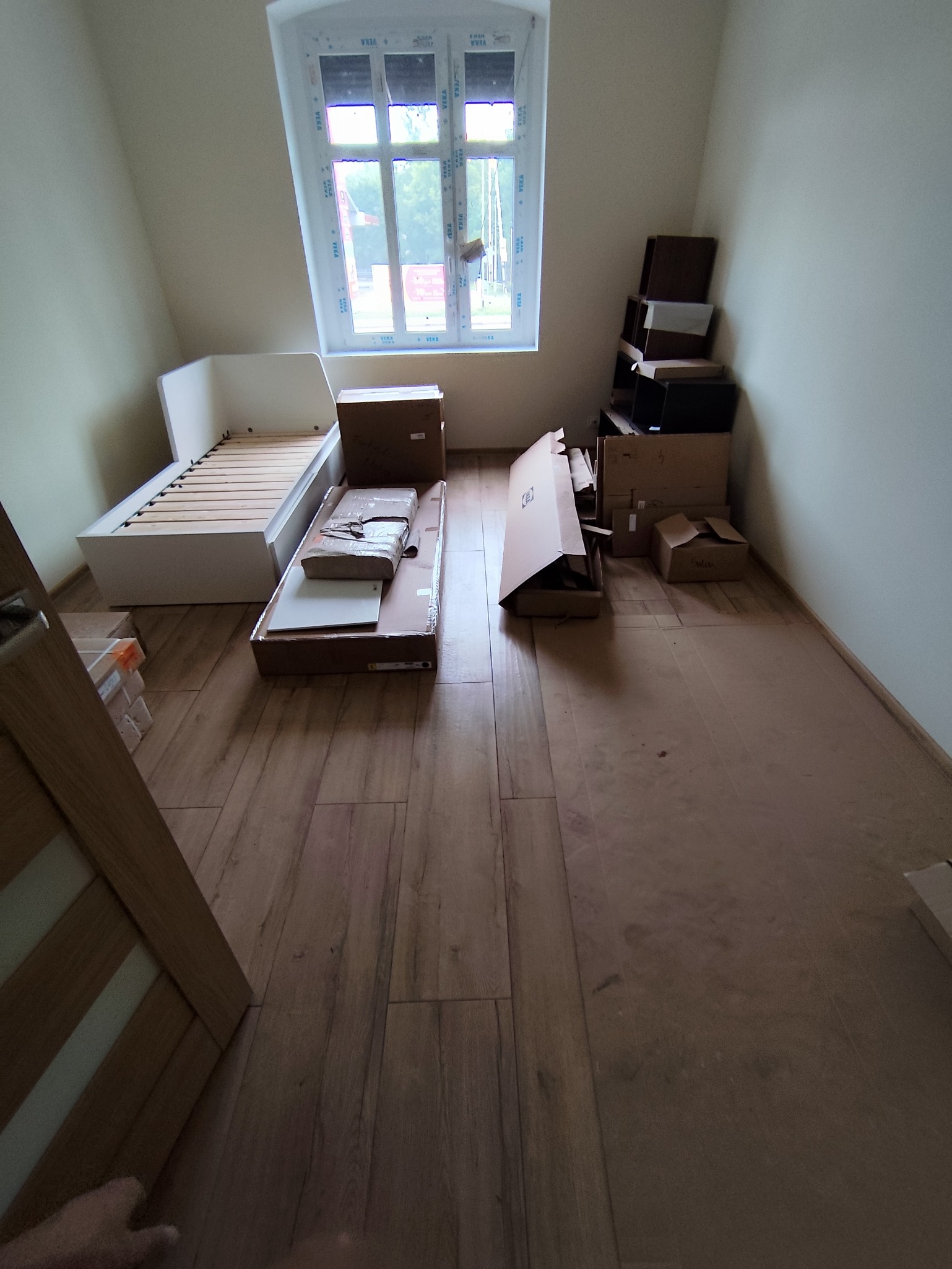 Poddasze z oknem dachowym, w trakcie prac wykończeniowych: rozpakowane kartony z meblami do sypialni, w tym łóżko i regał, przygotowane do montażu.