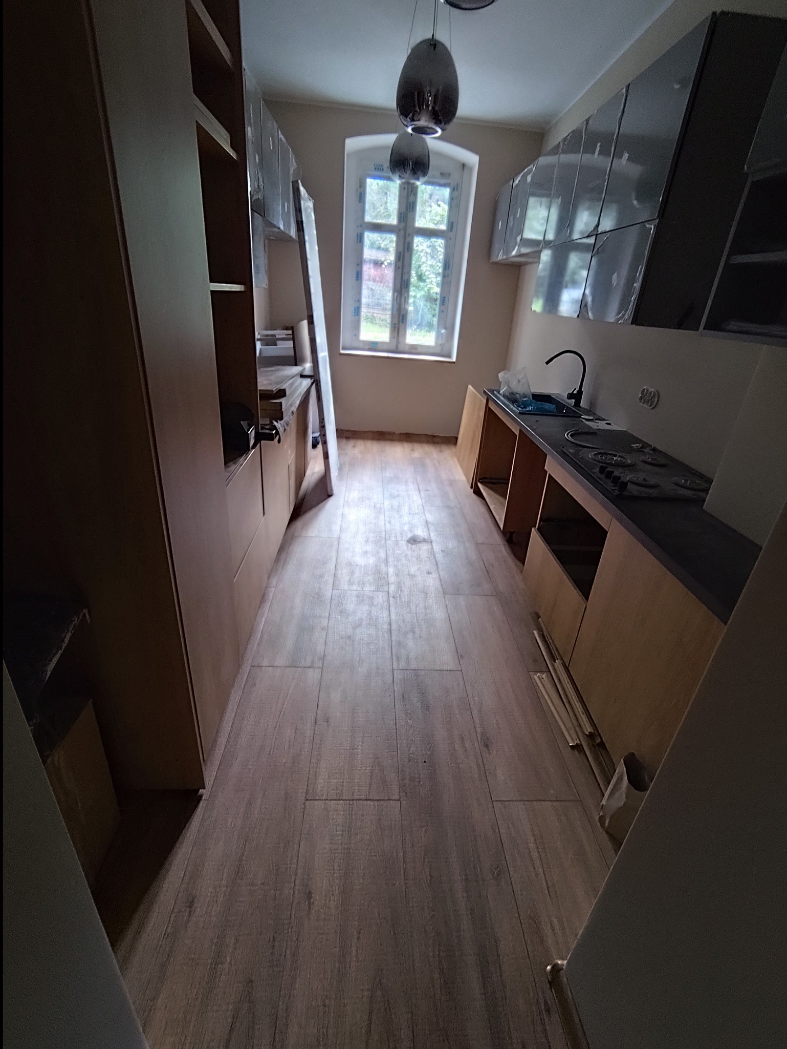 Wnętrze kuchni w trakcie montażu mebli: widoczne szafki wiszące w folii ochronnej, częściowo zamontowane szafki dolne z blatem, drewnopodobna podłoga i okno z widokiem na zieleń.