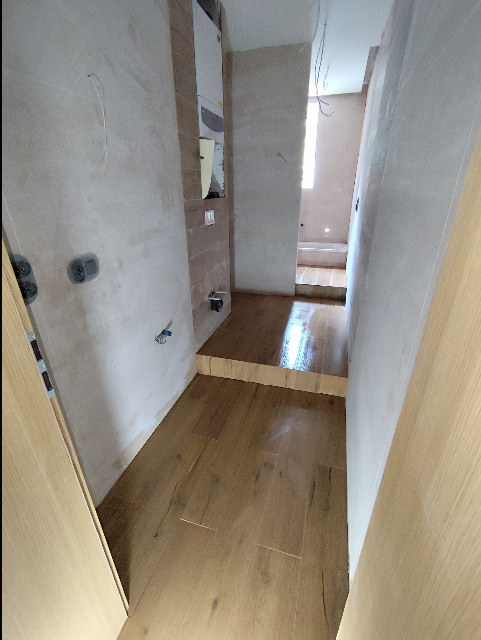 Remontowana łazienka z drewnopodobnymi płytkami na podłodze, niedokończone ściany z widocznymi puszkami elektrycznymi i rurami, etap prac wykończeniowych.