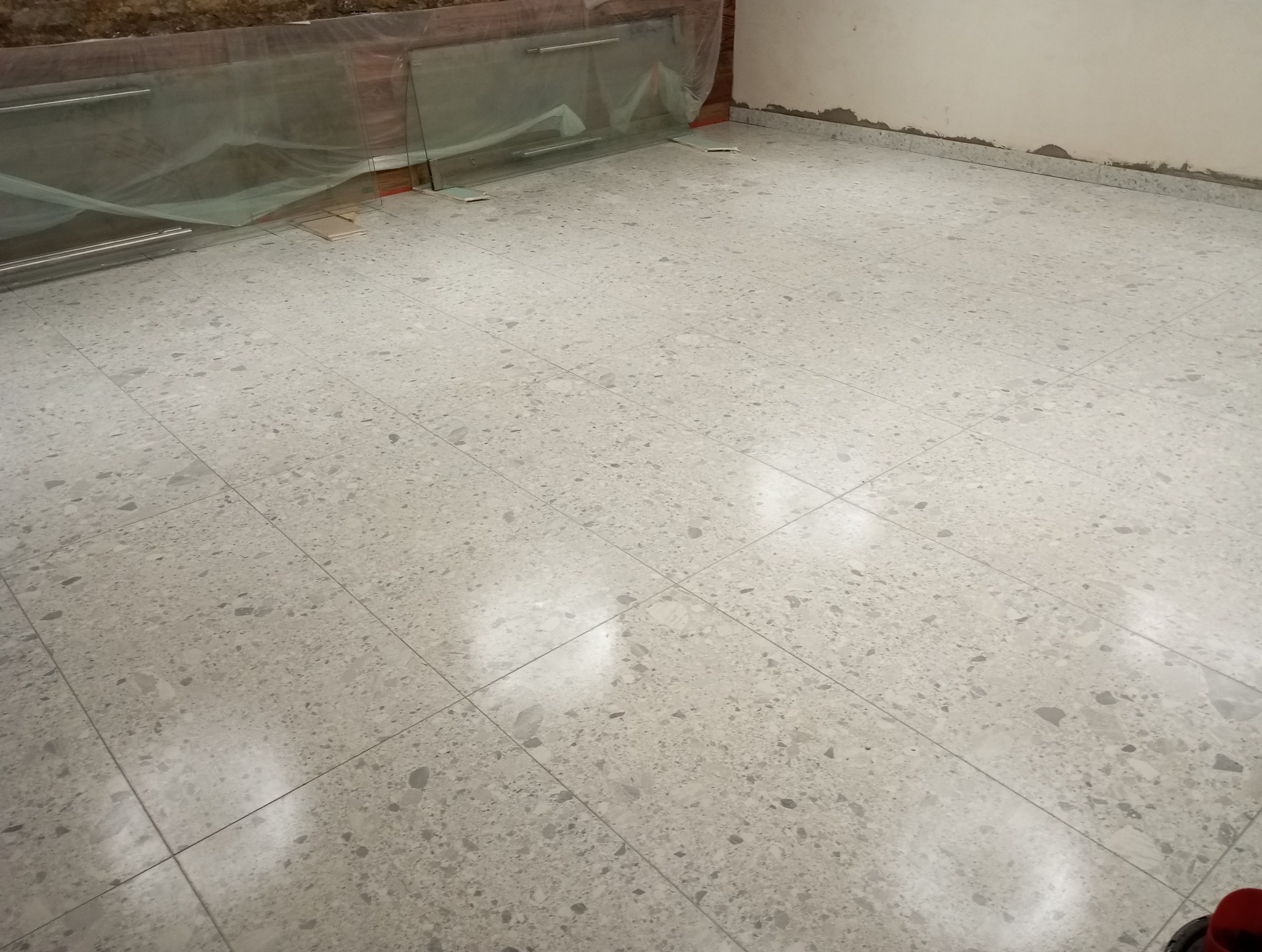 Nowa podłoga z terakoty w odcieniach szarości, z widocznymi płytami szklanymi zabezpieczonymi folią na ścianie, przygotowanymi do montażu.