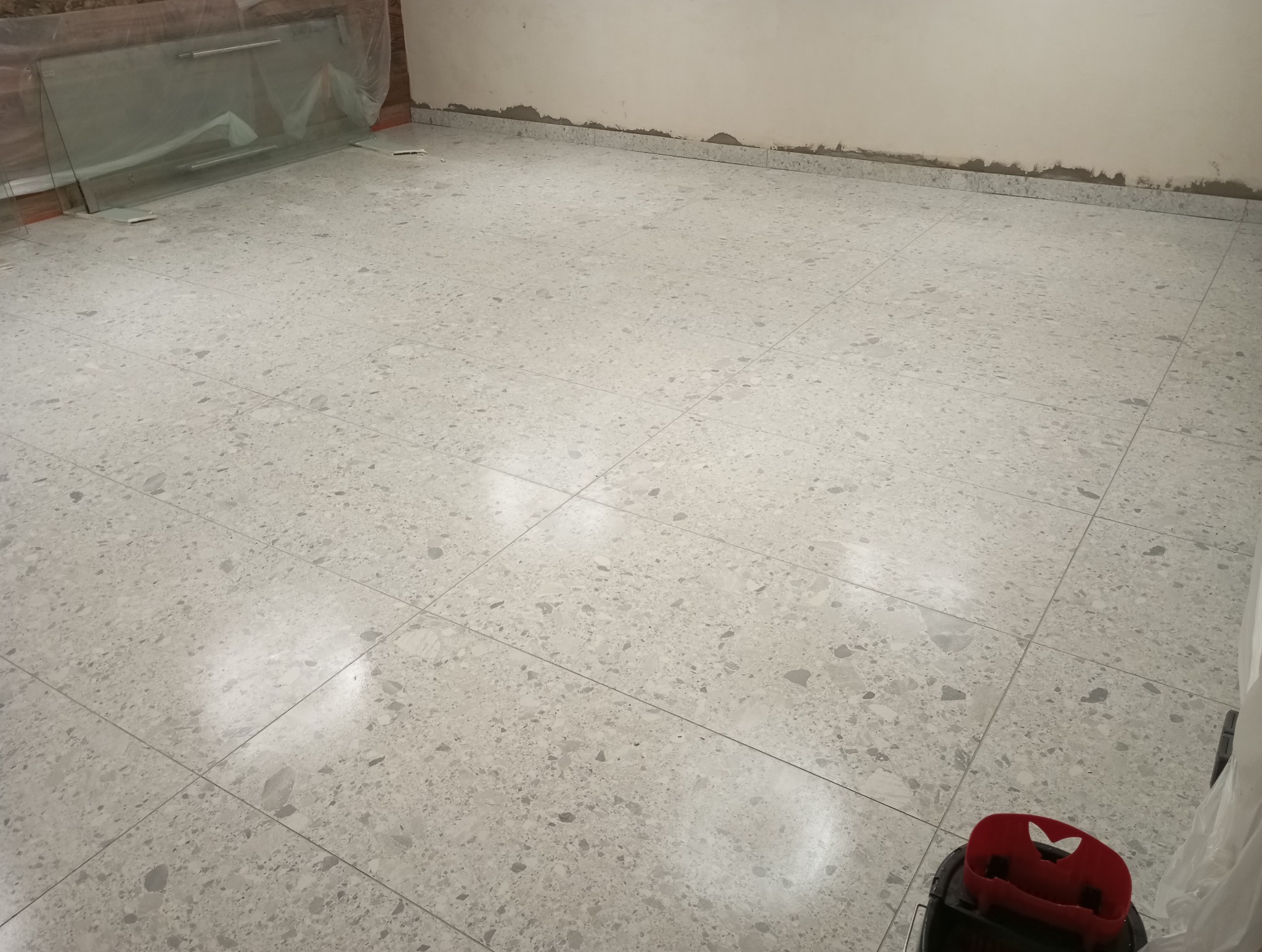 Świeżo położona, jasnoszara podłoga z płytek imitujących lastryko. Przy ścianach widoczne ślady po remoncie. Zabezpieczone szkło opiera się o ścianę. W rogu widoczny mop.