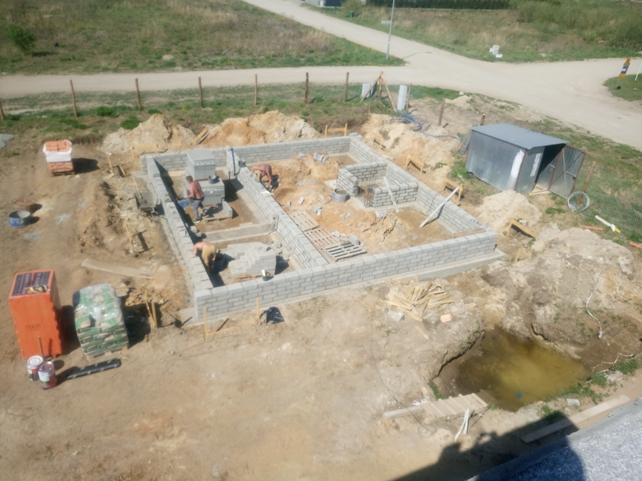 Budowa fundamentów domu z bloczków betonowych, widok z góry, dwóch murarzy przy pracy, palety z materiałami budowlanymi obok wykopu, na działce widoczna kałuża wody.