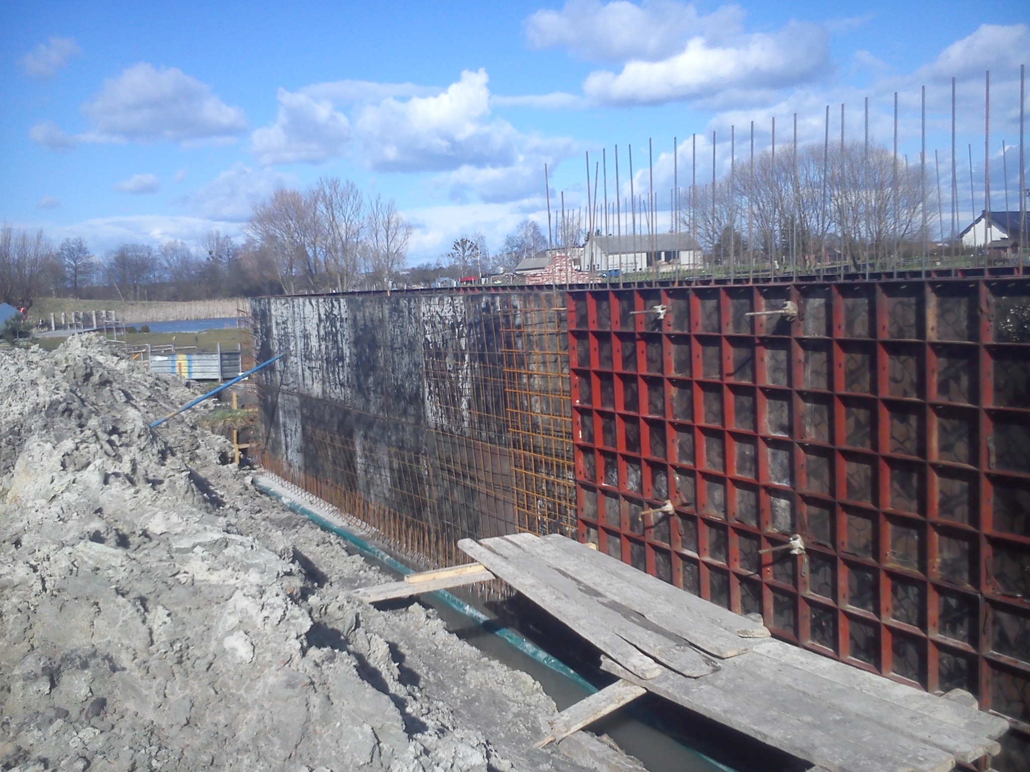 Szczegółowe ujęcie szalunku fundamentowego z widoczną konstrukcją stalową i deskami, przygotowanego do zalewania betonem, z hałdą ziemi w tle i fragmentem krajobrazu z jeziorem