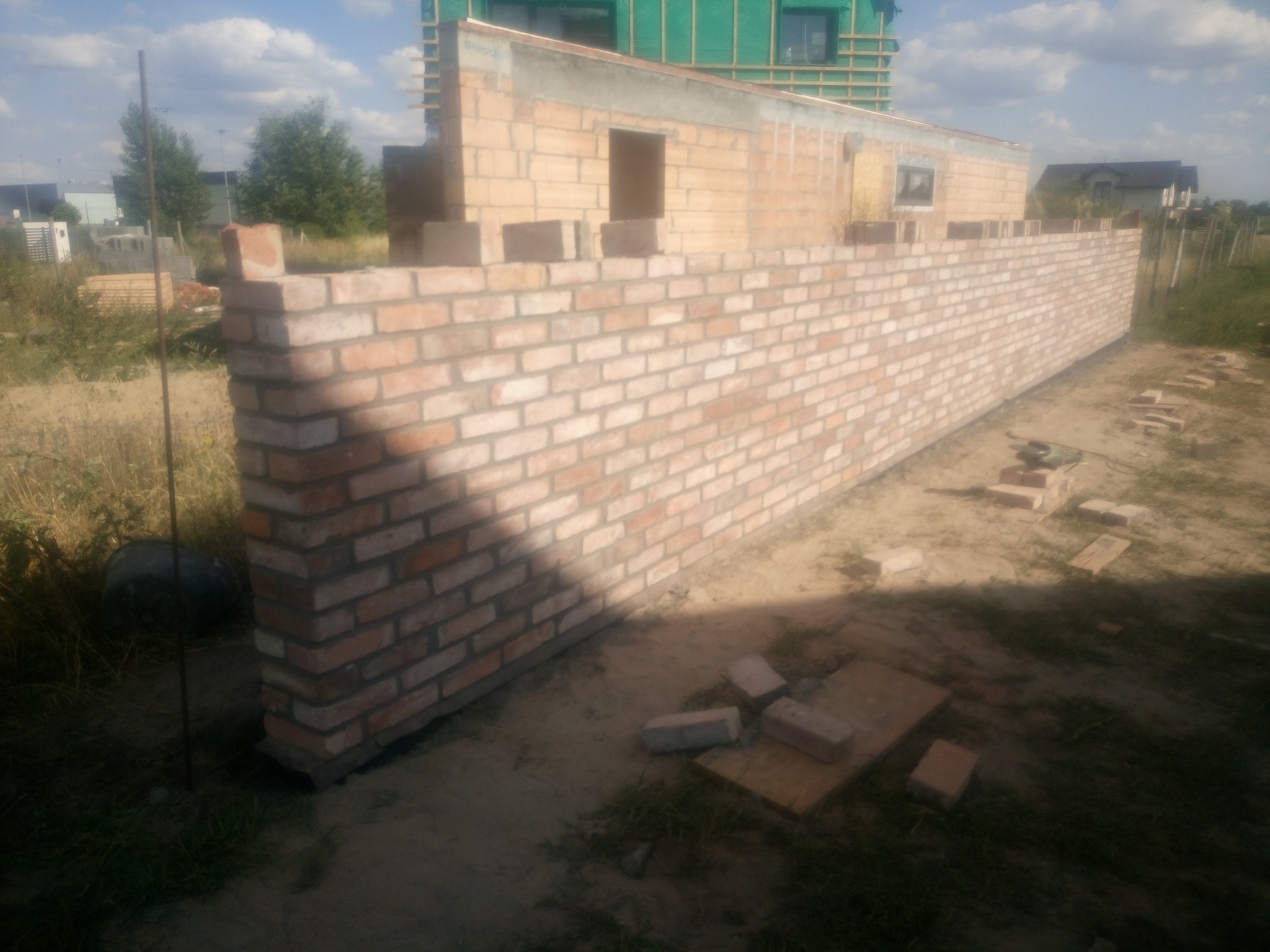 Wznoszenie muru z cegły klinkierowej w trakcie budowy domu jednorodzinnego, widoczne niezwiązane cegły i początki ścian wyższych kondygnacji.