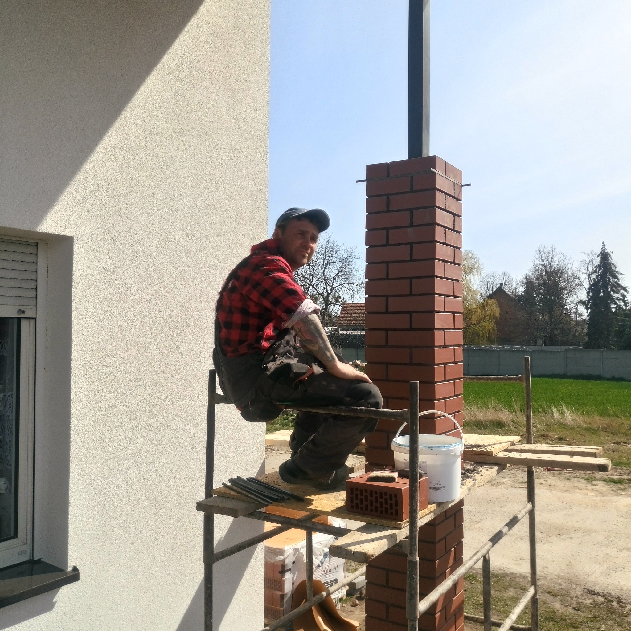 Murarz w czerwonej koszuli w kratę siedzi na rusztowaniu podczas wznoszenia ceglanego filaru przy ścianie domu, widoczne narzędzia i wiadro z zaprawą.