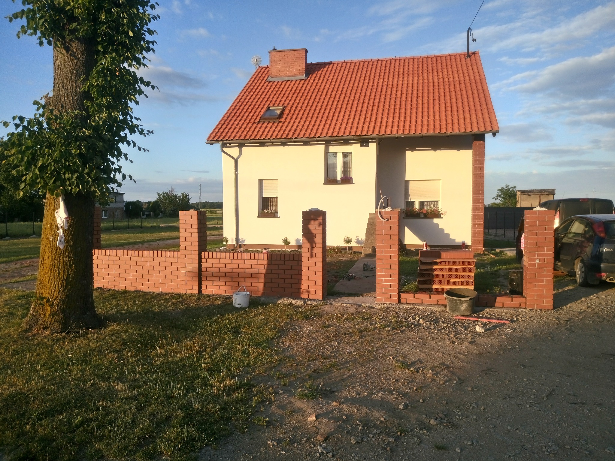 Niedawno wybudowany dom jednorodzinny z czerwonym dachem i częściowo wzniesionym ceglanym ogrodzeniem frontowym. Widoczne dwa wiadra budowlane i zaparkowany samochód.