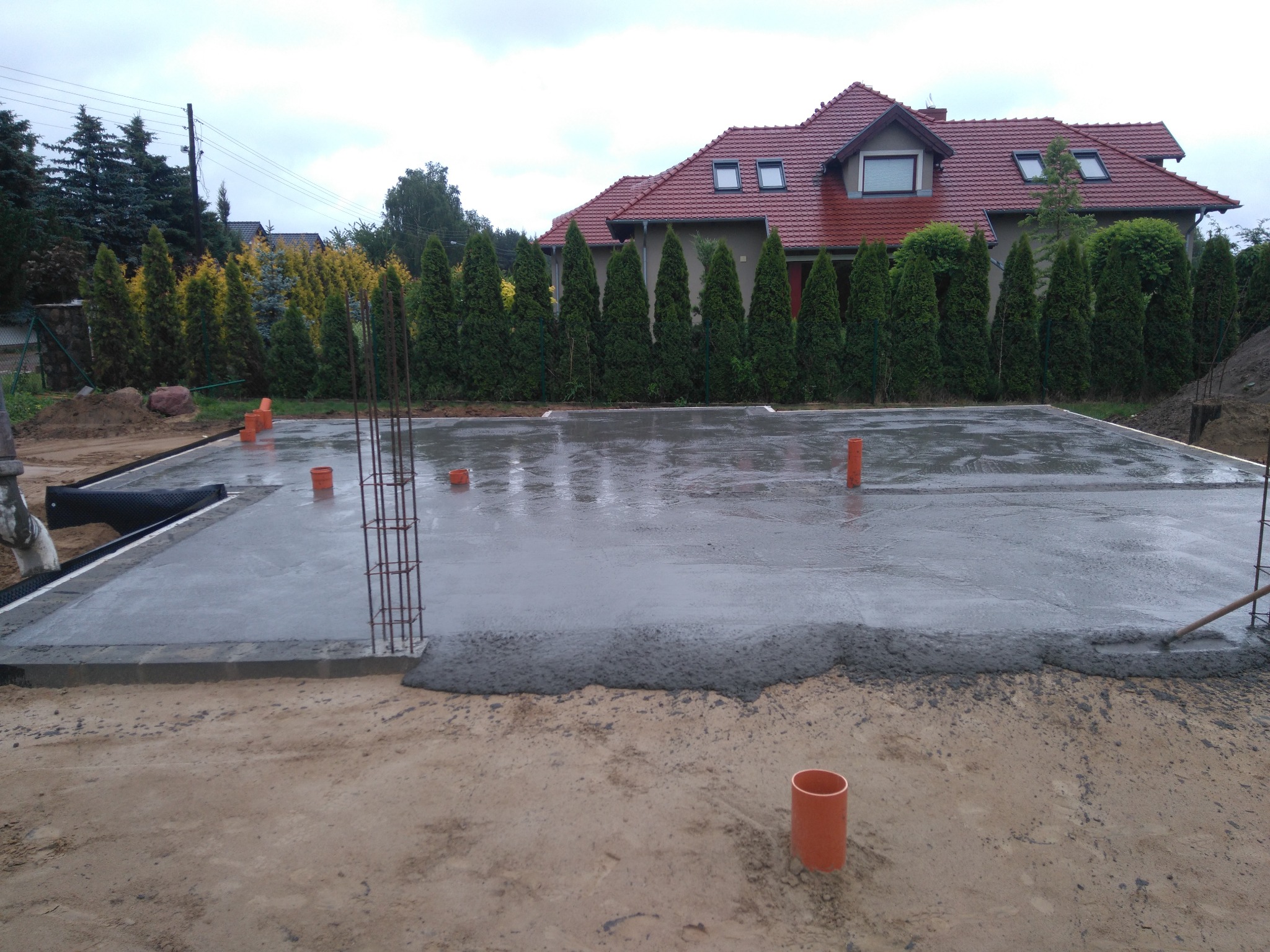 Wylany fundament pod budowę domu jednorodzinnego, widoczne zbrojenia i rury kanalizacyjne, w tle dom z czerwoną dachówką i zielone tuje.