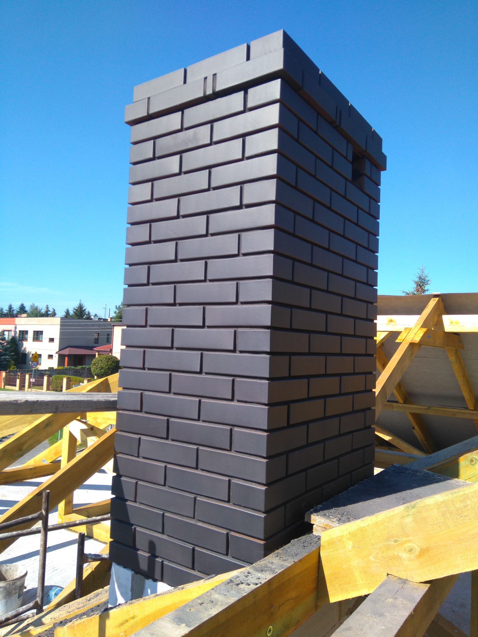 Wysoki komin z czarnej cegły klinkierowej, wystający ponad konstrukcję dachu w trakcie budowy, na tle błękitnego nieba.