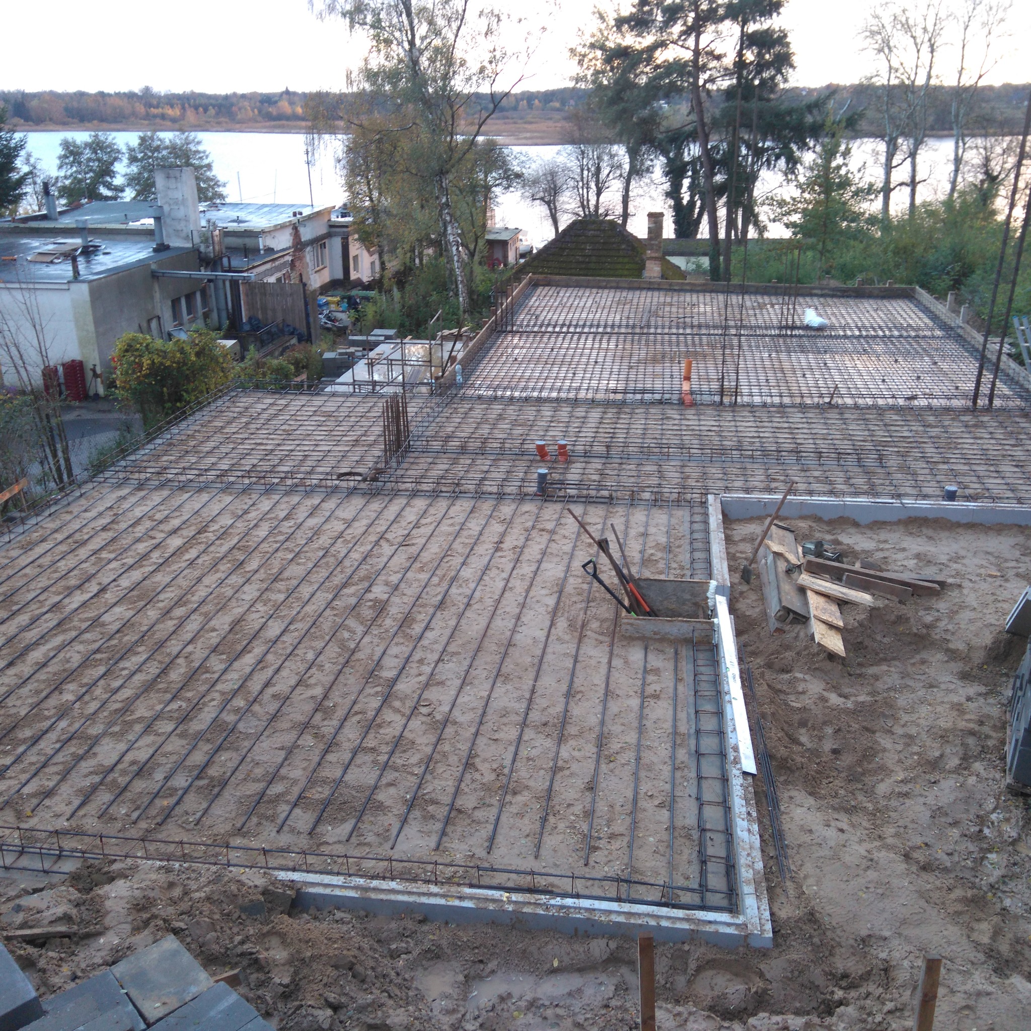 Zbrojenie płyty fundamentowej z widocznymi prętami, szalunkiem i narzędziami w wykopie, z oddalonym widokiem na jezioro i zabudowania.