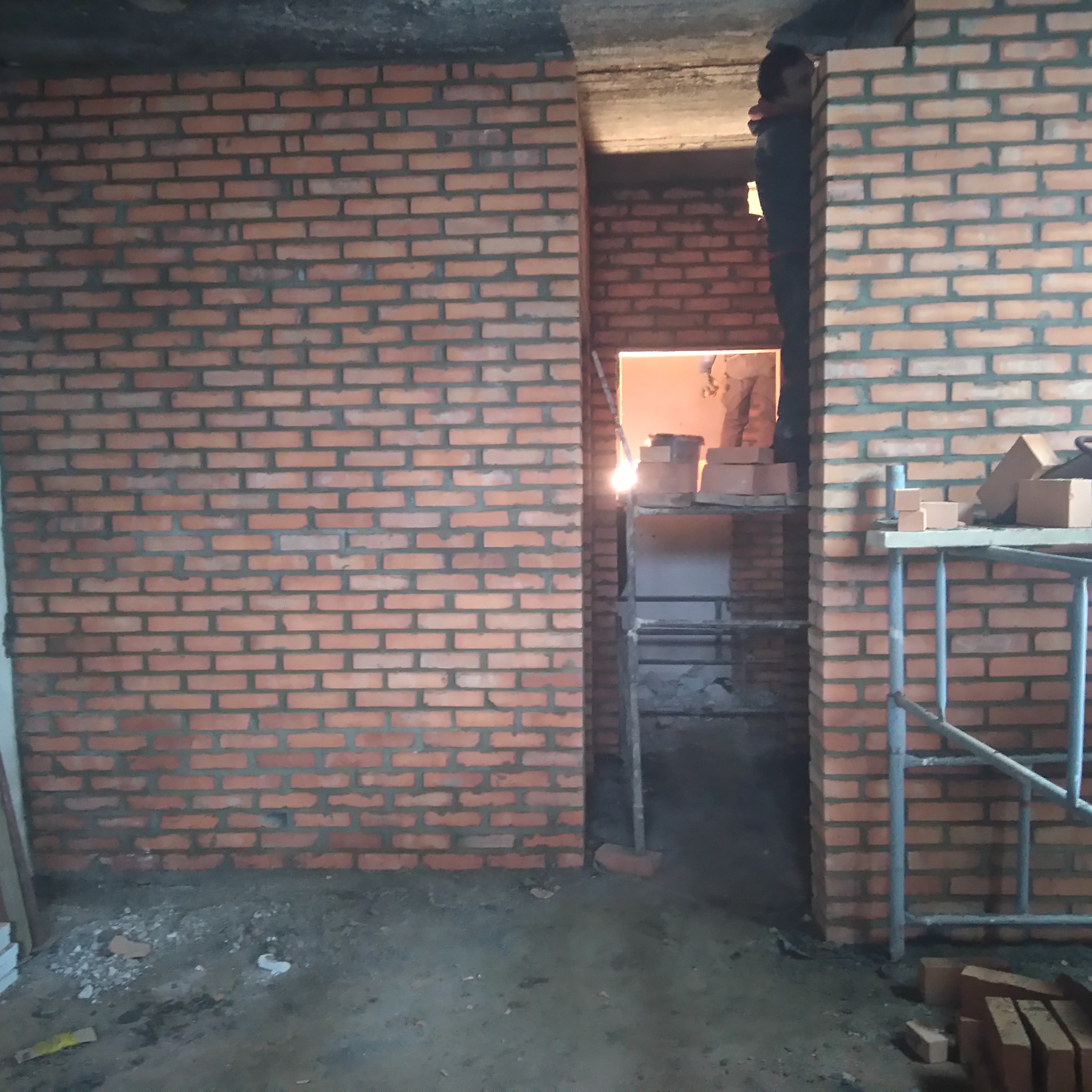 Wznoszenie ściany z czerwonej cegły, widoczne rusztowanie i zapas cegieł w prześwicie ściany, zarys sylwetki osoby pracującej na rusztowaniu.