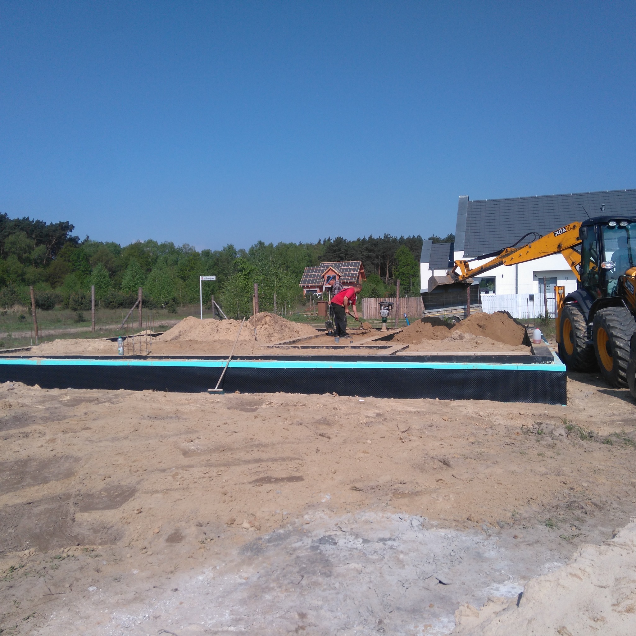 Przygotowanie fundamentów pod budowę domu: izolacja pionowa, wykop, pracownik z łopatą i koparka w tle.