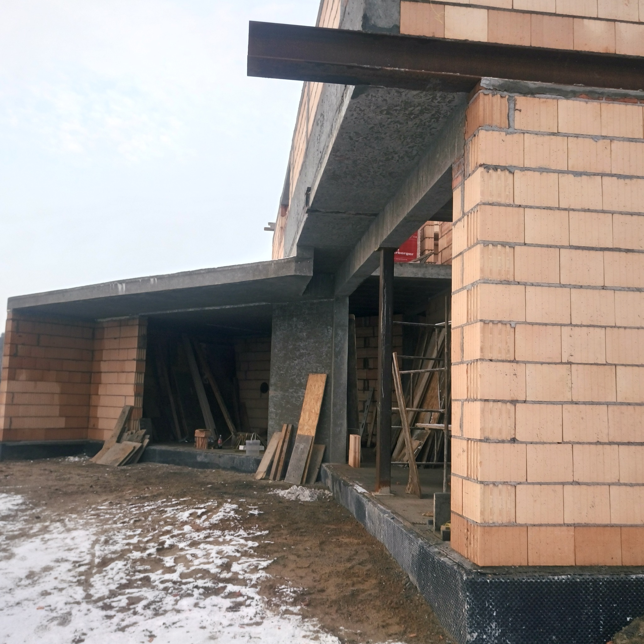 Surowy stan domu w trakcie budowy, widoczne ściany z pustaków ceramicznych, betonowy strop, stalowa belka nad otworem, częściowo ośnieżony teren budowy.
