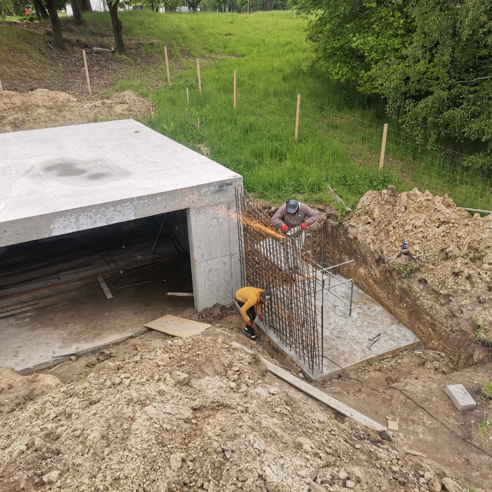 Prace zbrojarskie przy budowie fundamentów: dwóch pracowników tnie pręty zbrojeniowe szlifierką kątową, iskry lecą spod tarczy, widoczny betonowy strop nad przyszłym pomieszczeniem.