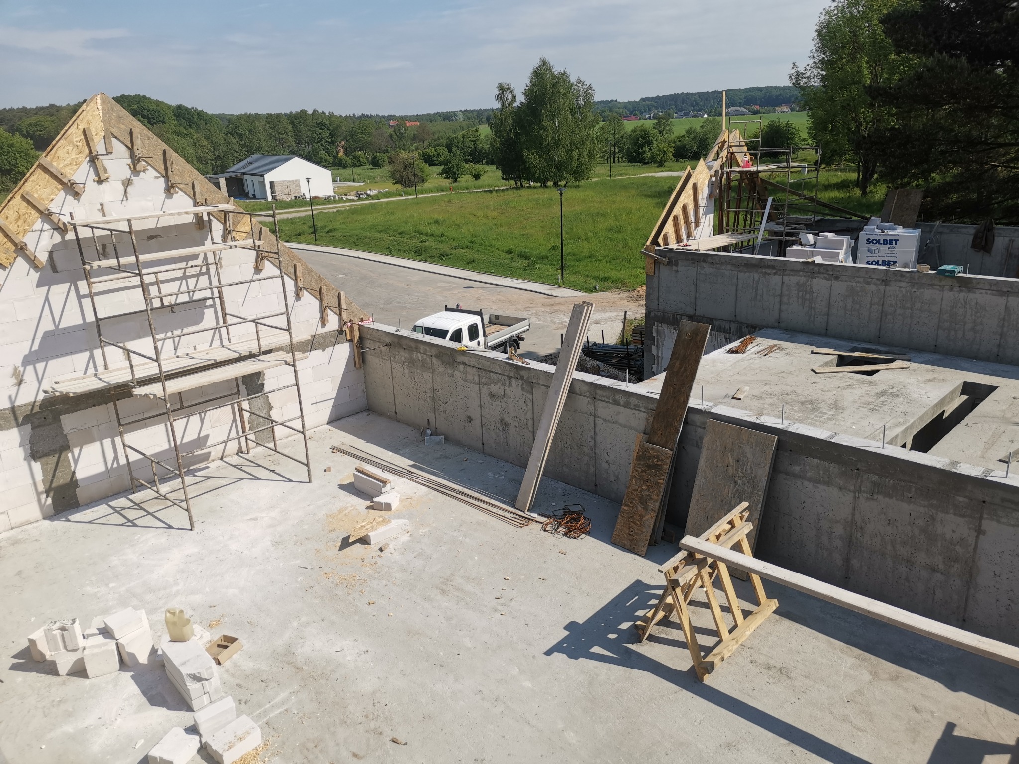 Budowa domu w stanie surowym otwartym: wzniesione ściany z bloczków, widoczne rusztowania, drewniana konstrukcja dachu w trakcie montażu, na podłożu leżą bloczki Solbet i narzędzia, w tle krajobraz.