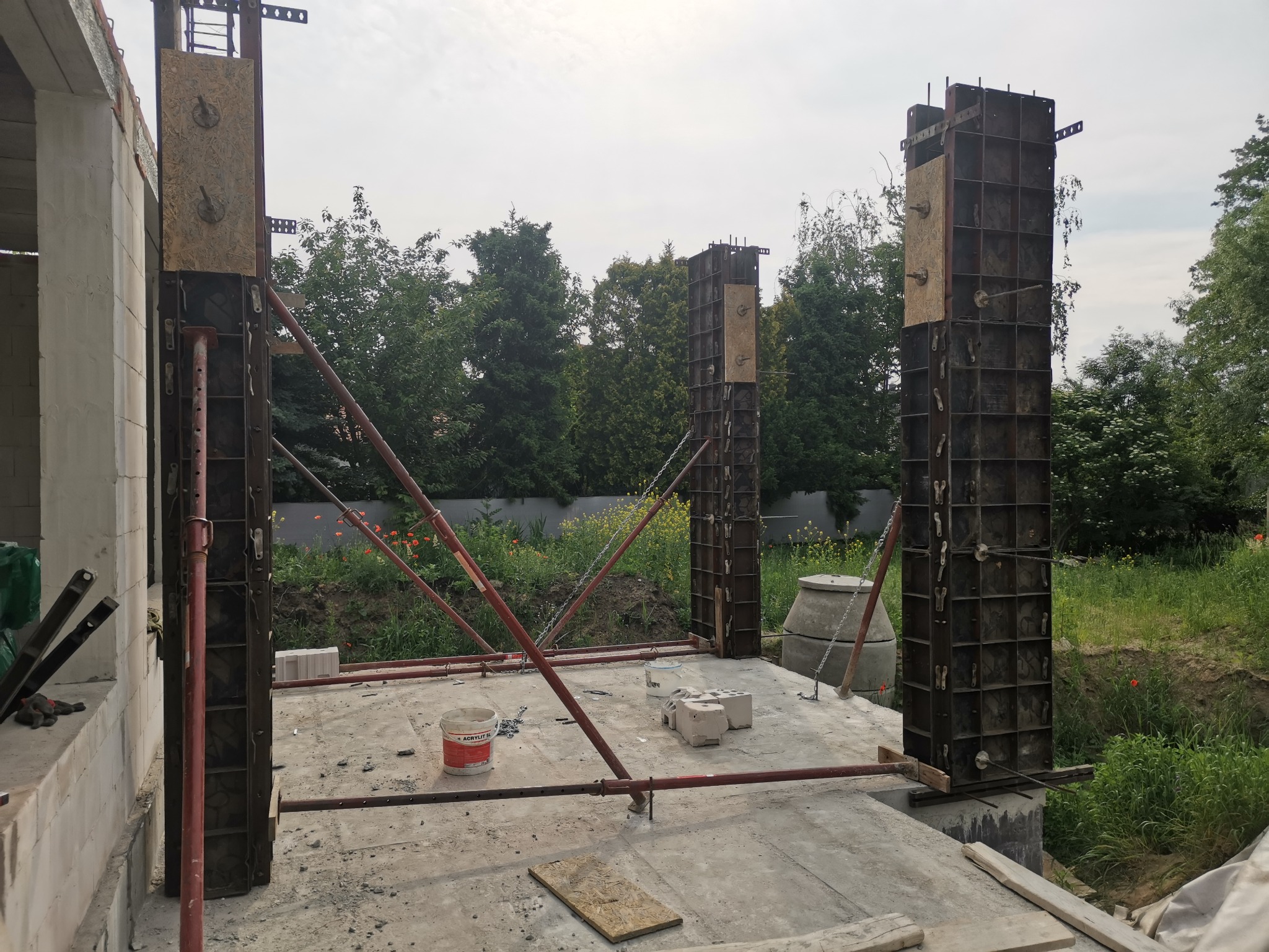 Wznoszenie szkieletu kolumn nośnych z użyciem szalunków stalowych i podpór teleskopowych na wylewce fundamentowej, widoczne materiały budowlane i otoczenie zieleni.