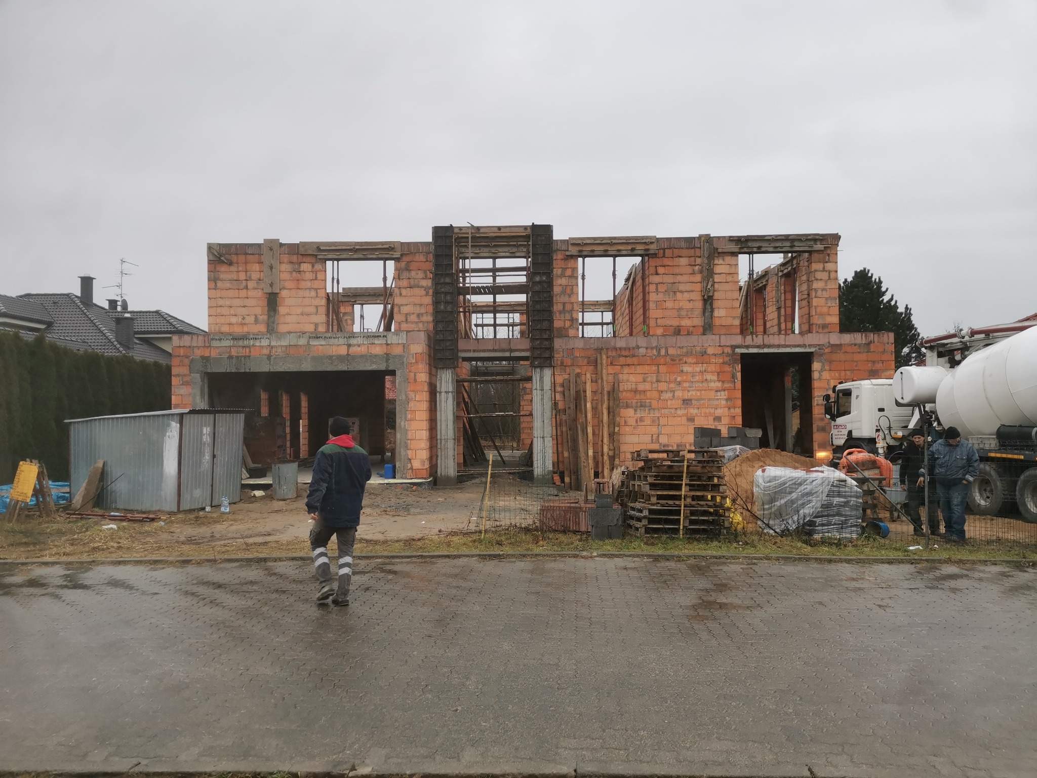 Budowa domu murowanego w stanie surowym otwartym; widoczny mur z czerwonej cegły, betonowy fundament, szalunki okienne, palety z materiałami budowlanymi oraz betoniarka w tle; pracownik budowlany...