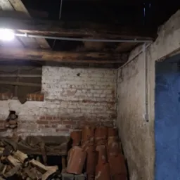 Wnętrze starego budynku z odsłoniętą cegłą, oświetlone lampą. Widoczne przewody elektryczne i elementy instalacji. Część pomieszczenia zagracona drewnem i dachówkami.